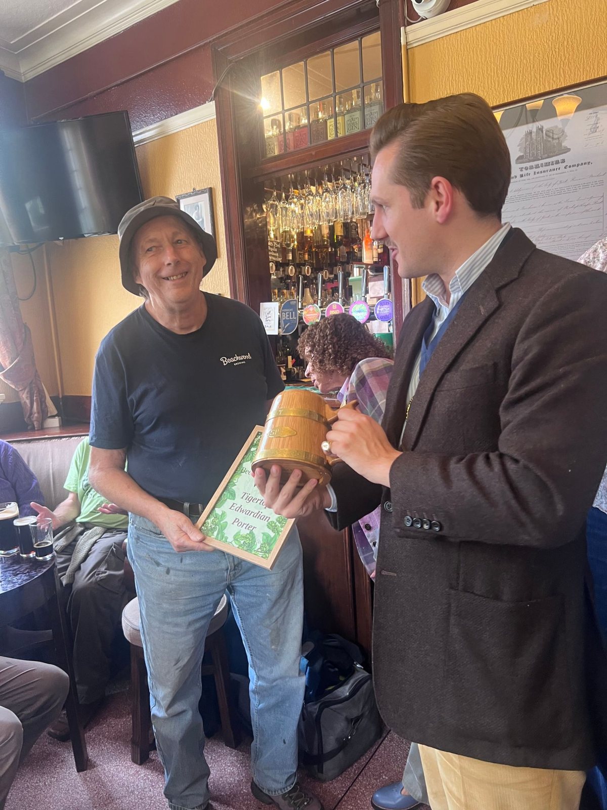 In the bar of the Swan, Liam Randall, in a brown tweed jacket, hands a small oak tankard bound with brass hoops to the smiling winner, who holds a framed certificate reading Tigertops Edwardian Porter.
