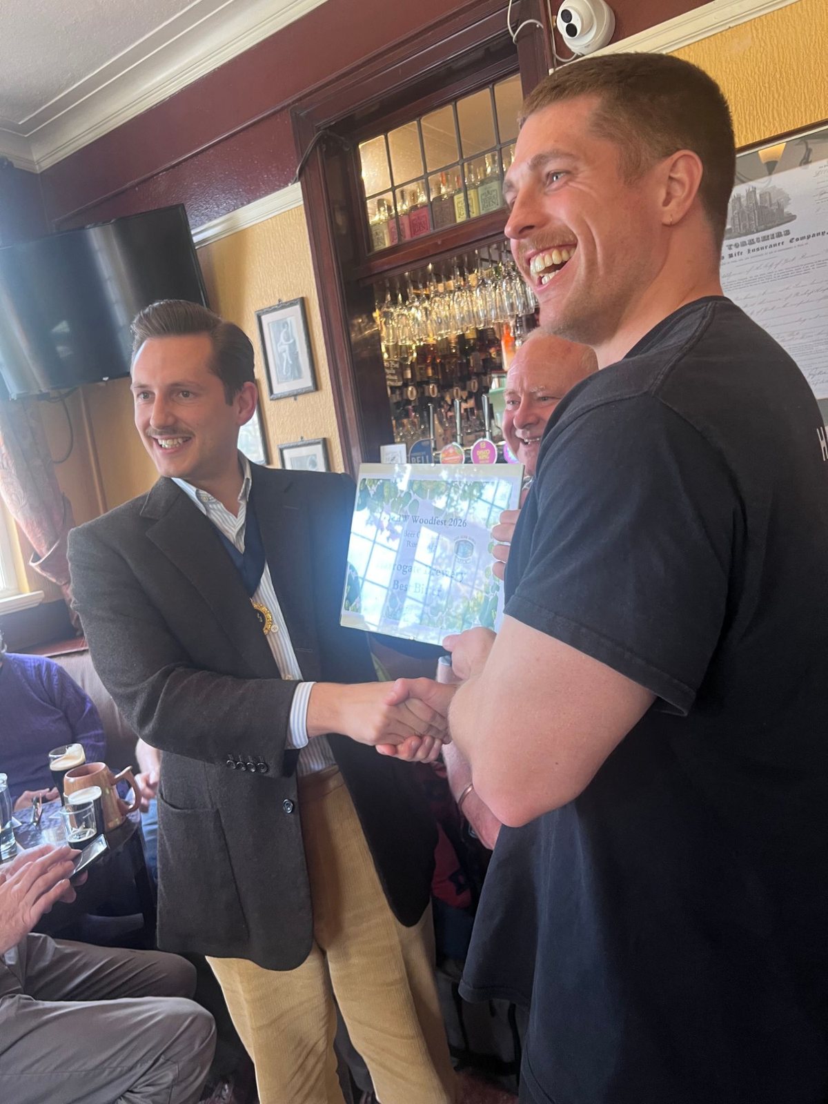 Liam Randall shakes hands with a smiling representative of Harrogate Brewery, who holds a framed Woodfest 2026 certificate for Harrogate Brewery Best Bitter, in the bar of the Swan.