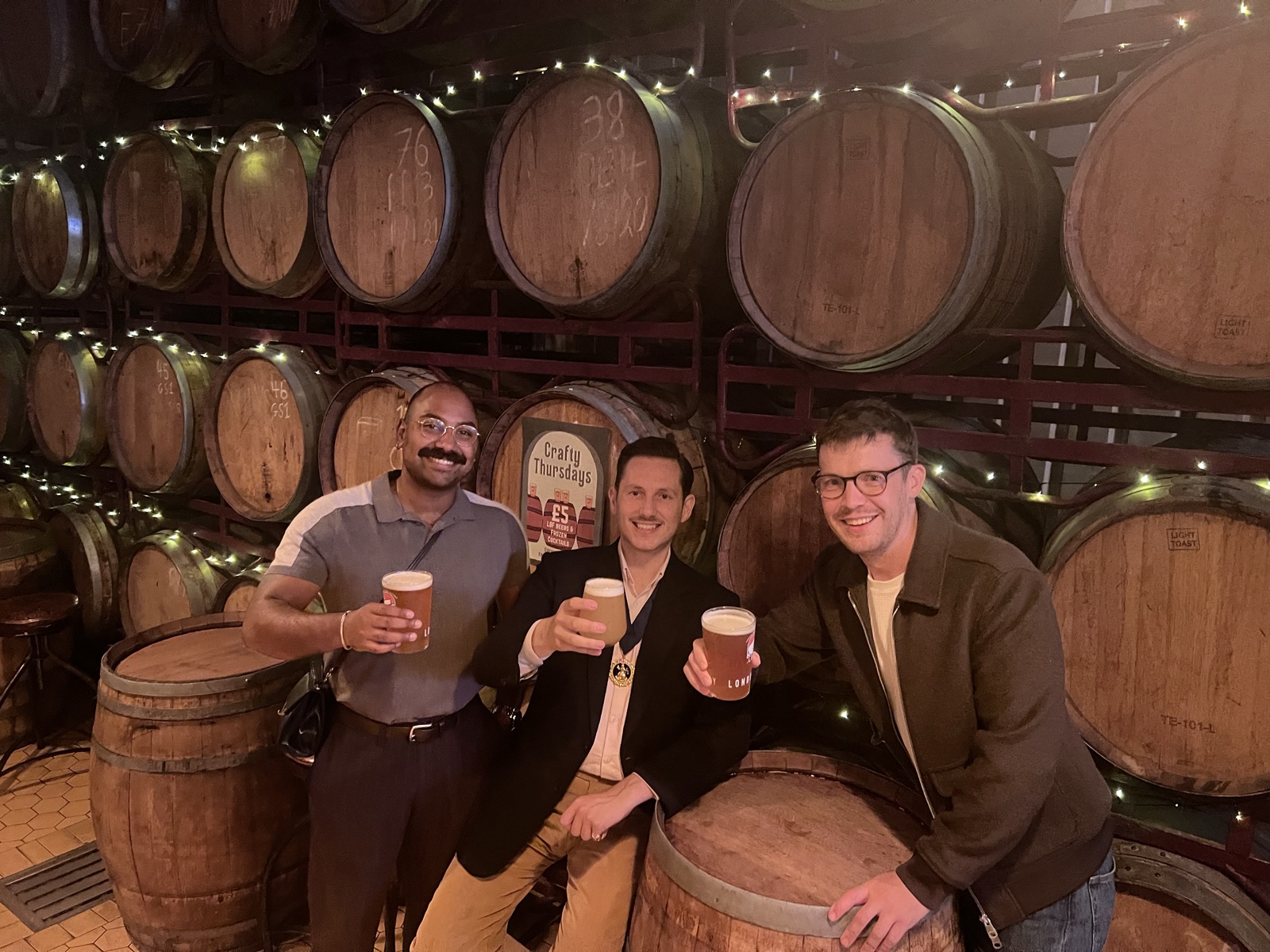 Three men raise pints in a railway-arch bar on the Bermondsey Beer Mile, before a wall of oak barrels stacked on racks and strung with fairy lights.