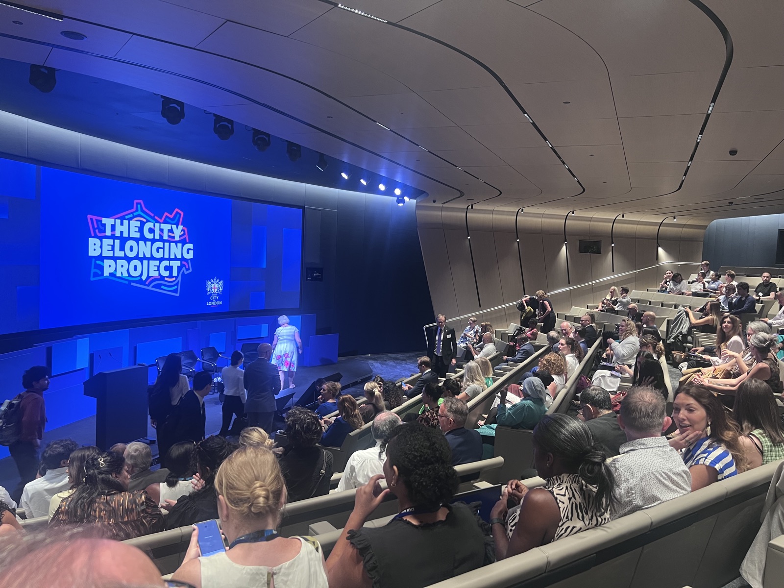 The curved, tiered auditorium at Deutsche Bank&rsquo;s Moorfields headquarters fills for the City Belonging Champions launch, its wide blue screen carrying the City Belonging Project logo beside the City of London Corporation&rsquo;s crest as guests find their seats.