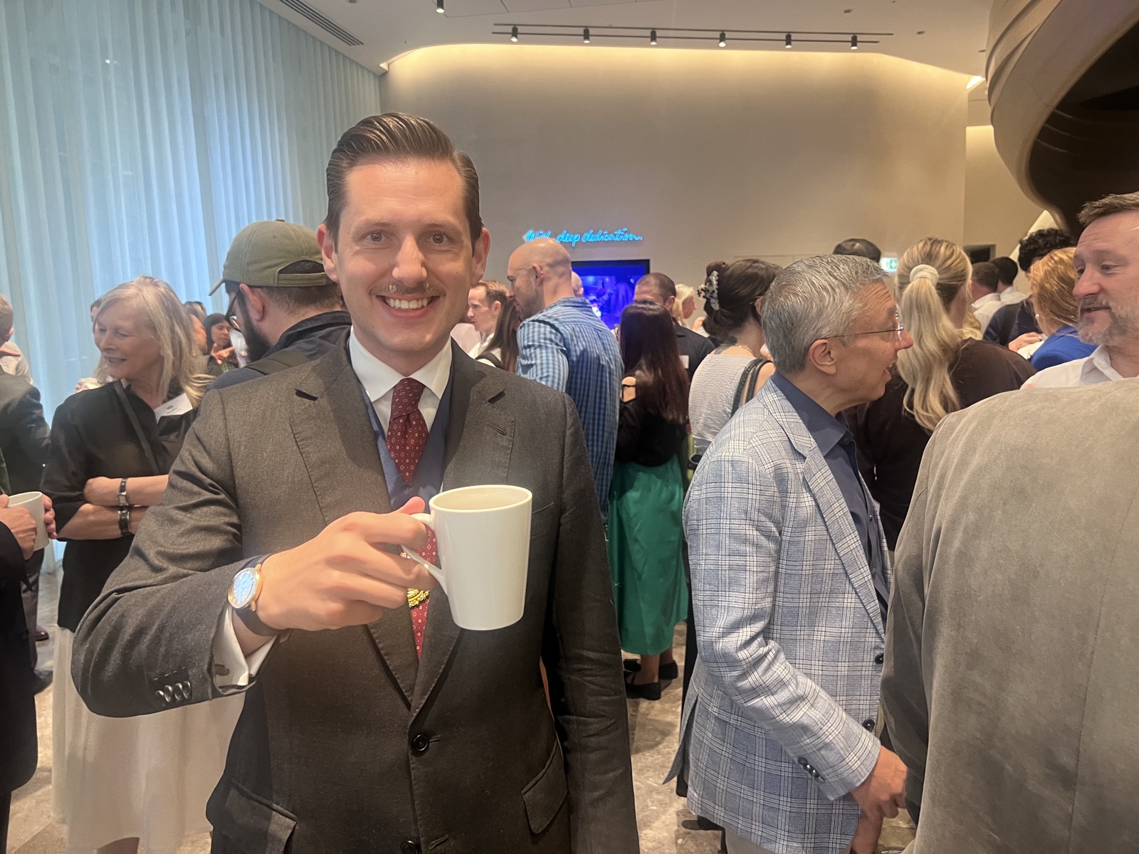 In a crowded reception space at Deutsche Bank, Liam Randall in a grey three-piece suit with a navy waistcoat and burgundy patterned tie smiles at the camera and raises a white mug of coffee; behind him guests mingle beneath a blue neon sign and floor-to-ceiling curtains.