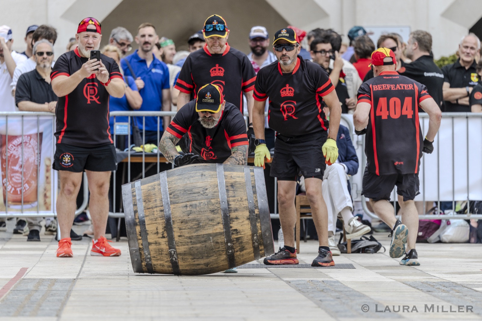 Mid-race, a team of Yeoman Warders in black-and-red kit bearing the crowned royal cypher steers a dark cask through the turn, one racer crouched low over the barrel and a teammate in a shirt reading BEEFEATER 404 sprinting behind, spectators lining the barriers of Guildhall Yard.
