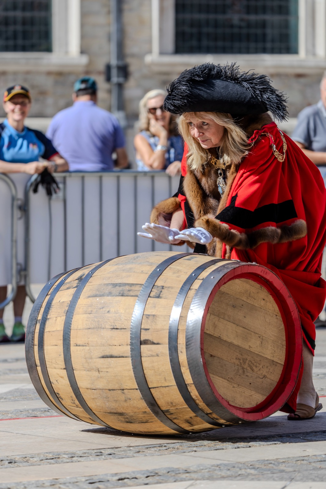 The Lady Mayor, Dame Susan Langley, in scarlet robes trimmed with fur, gold chains of office, white gloves and a black plumed tricorn hat, bends to roll a pale new oak cask with a red-painted head across the stone paving of Guildhall Yard, spectators watching from behind a barrier.