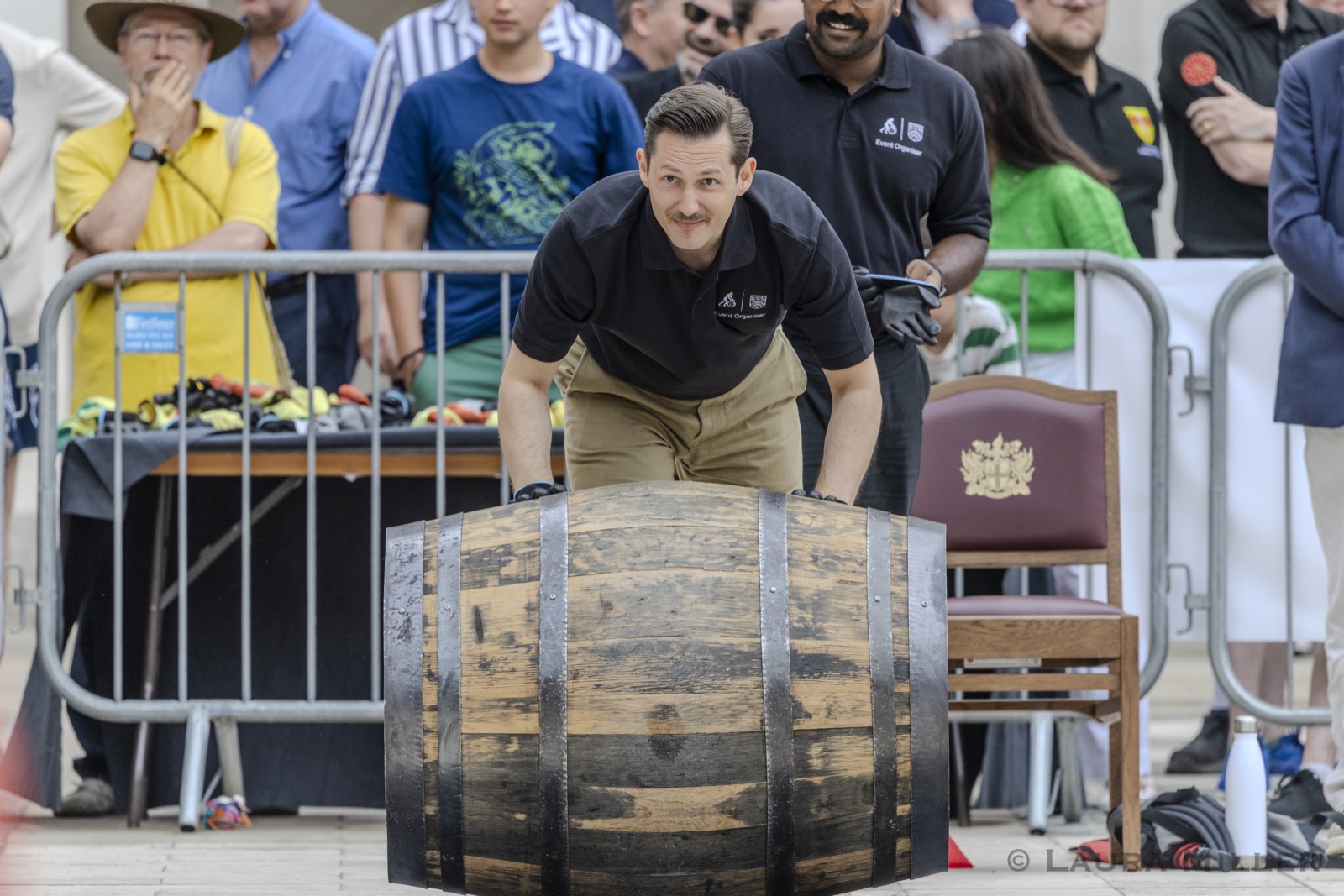 Liam Randall, in a black Event Organiser polo shirt and gloves, crouches ready to roll a dark charred cask at the start line, spectators at the barriers behind him and a ceremonial chair embroidered with the City of London arms to one side.