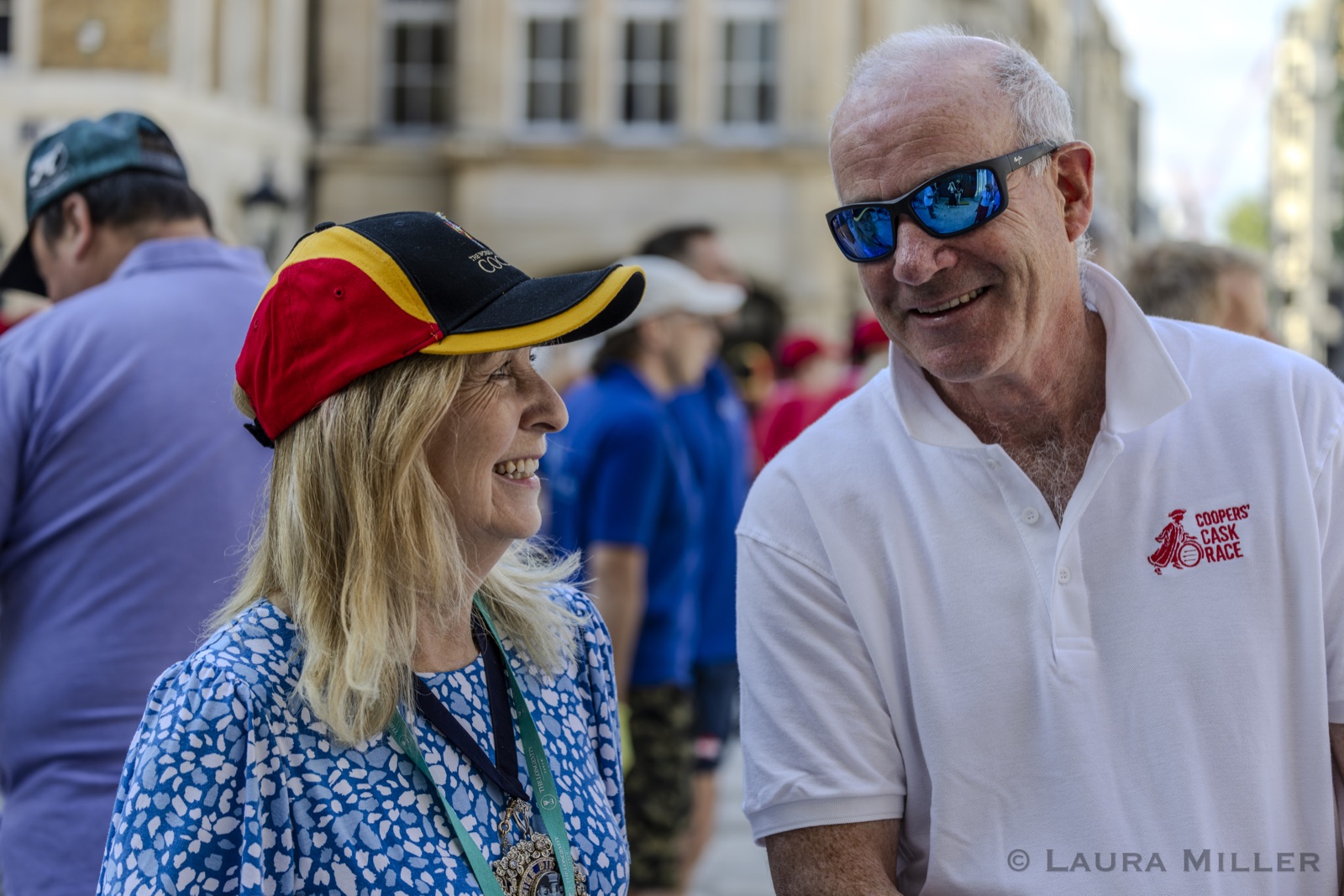 The Lady Mayor, in a Coopers&rsquo; cap of red, yellow and black with her badge on a lanyard, laughs with a member of the mayoral team wearing the white custom-embroidered Coopers&rsquo; Cask Race polo shirt, racers milling in the sunshine behind them.