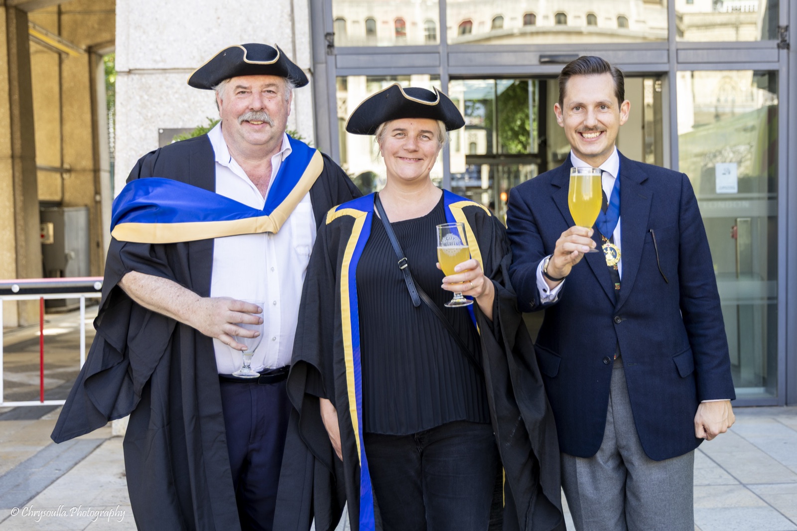 Three Ale Conners raise glasses of golden festival beer outside the Guildhall complex: two fellow conners in tricorn hats and black gowns faced with blue and gold, and Liam Randall on the right in a navy blazer, his badge of office on a ribbon round his neck.