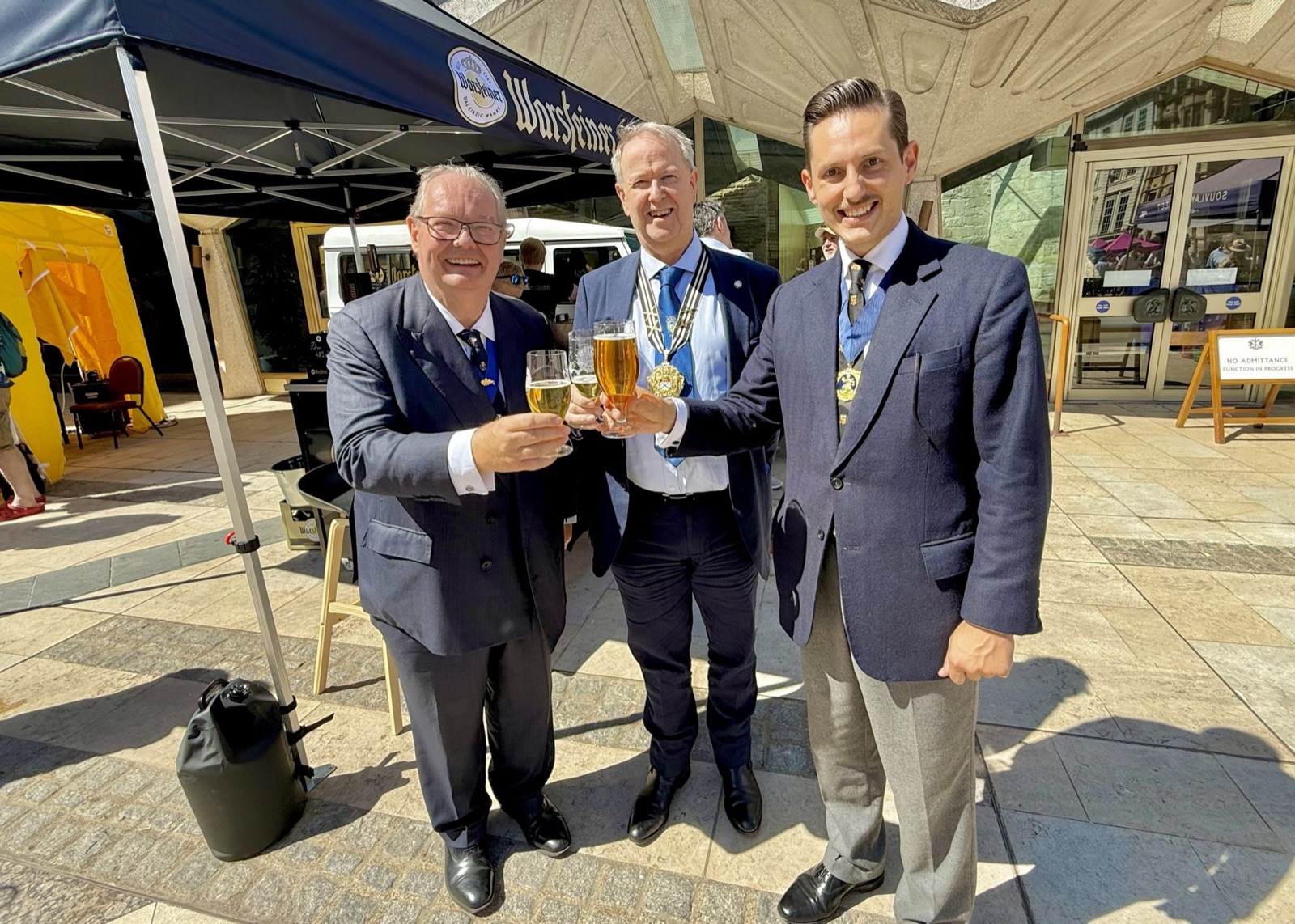 Three men clink glasses of beer in a toast beneath a festival gazebo in Guildhall Yard on a sunny afternoon; Liam Randall, wearing a livery badge, is on the right.