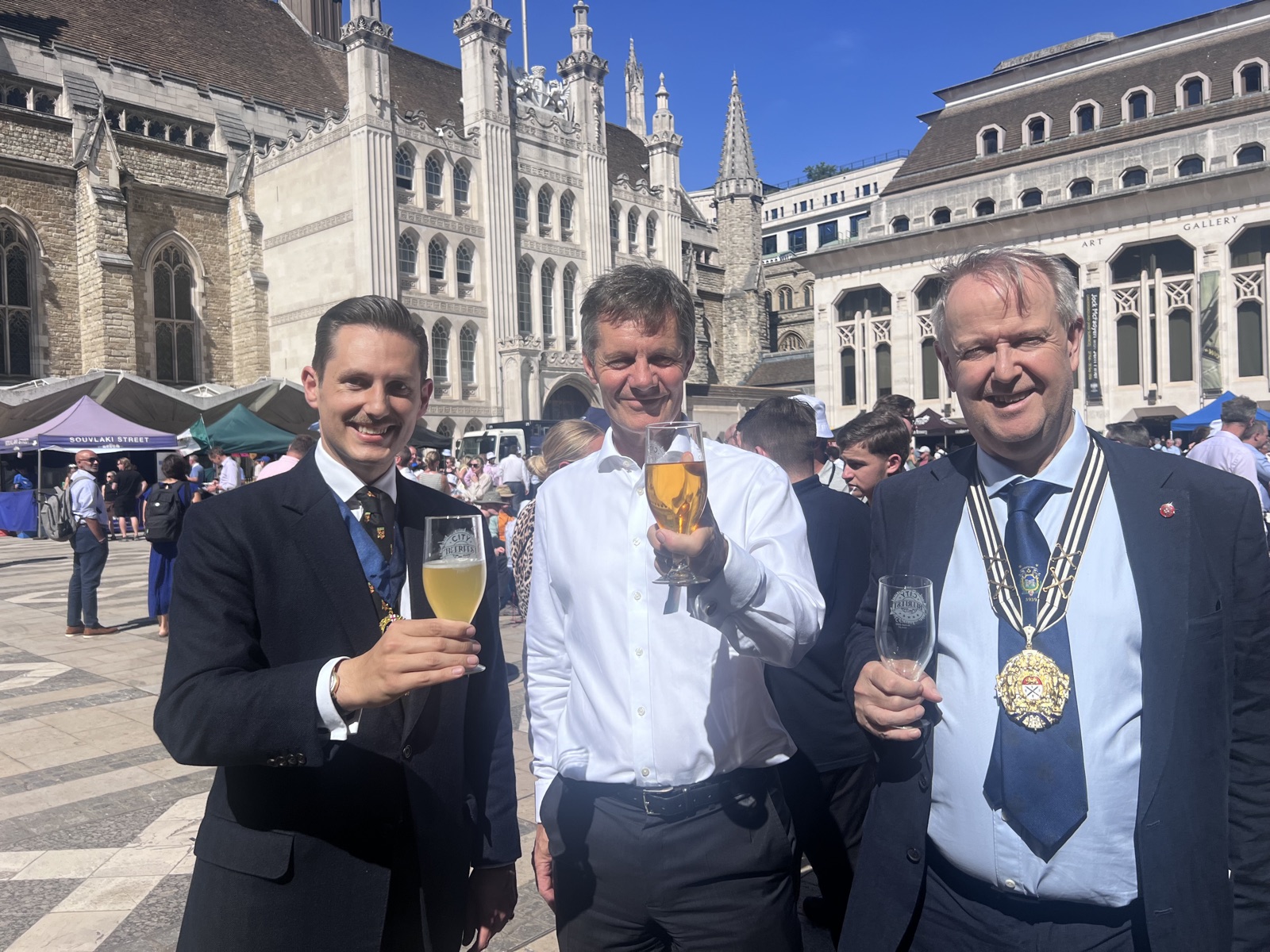 Three men raise glasses of City Beer Festival ale in Guildhall Yard, the Gothic front of Guildhall and the Guildhall Art Gallery behind them under a clear blue sky; Liam Randall stands on the left.