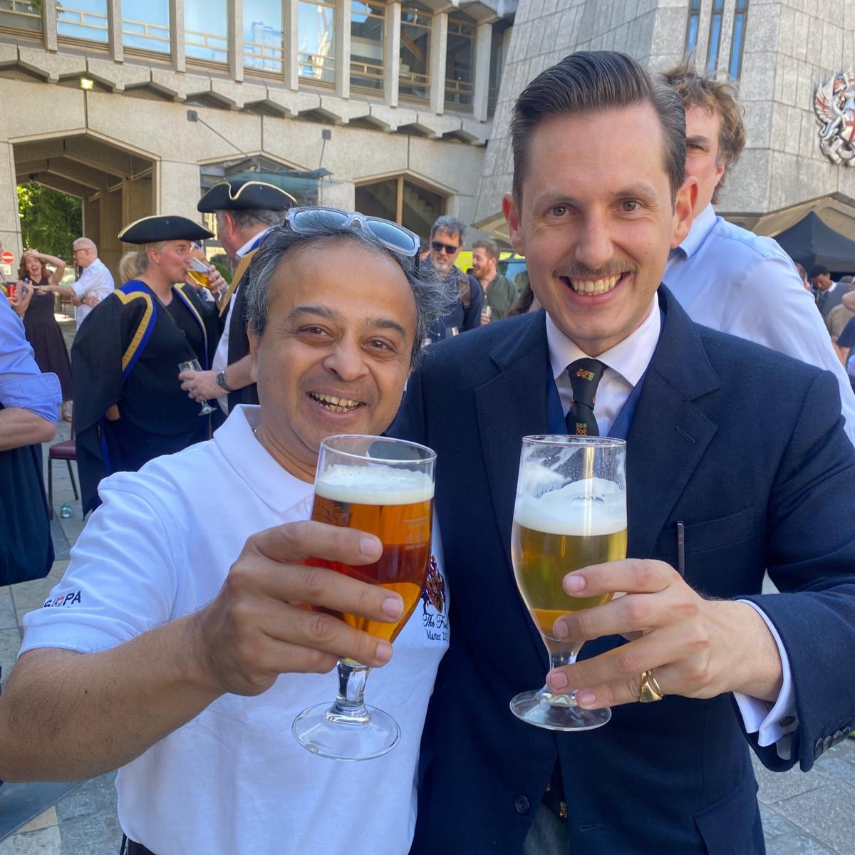 Liam Randall, in a dark suit, and Ashutosh Shastri, Master of the Fuellers&rsquo; Company, in an embroidered white polo shirt, raise glasses of ale in a toast at the City Beer Festival, with fellow guests and a City official in a tricorn hat behind them.