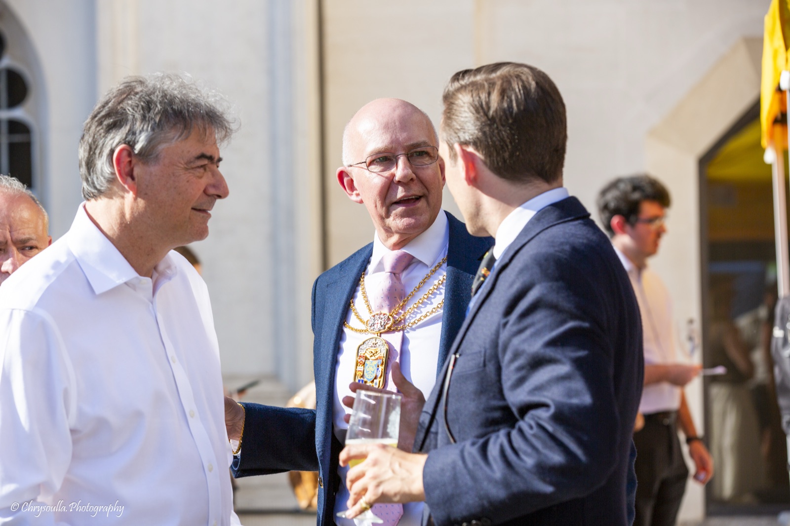 Keith Bottomley, the Non-Aldermanic Sheriff, in his chains and badge of office with a glass of festival ale in hand, in sunny conversation with a guest in a white shirt and with Liam Randall, seen from the side on the right.