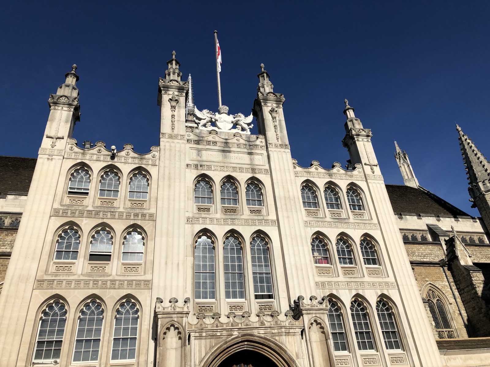 The Grand Entrance of Guildhall in bright sunshine beneath a deep blue sky: a Portland-stone porch crowned by the City of London&rsquo;s coat of arms between four pinnacles, the City flag flying above and the older stonework of the Great Hall rising to the right.