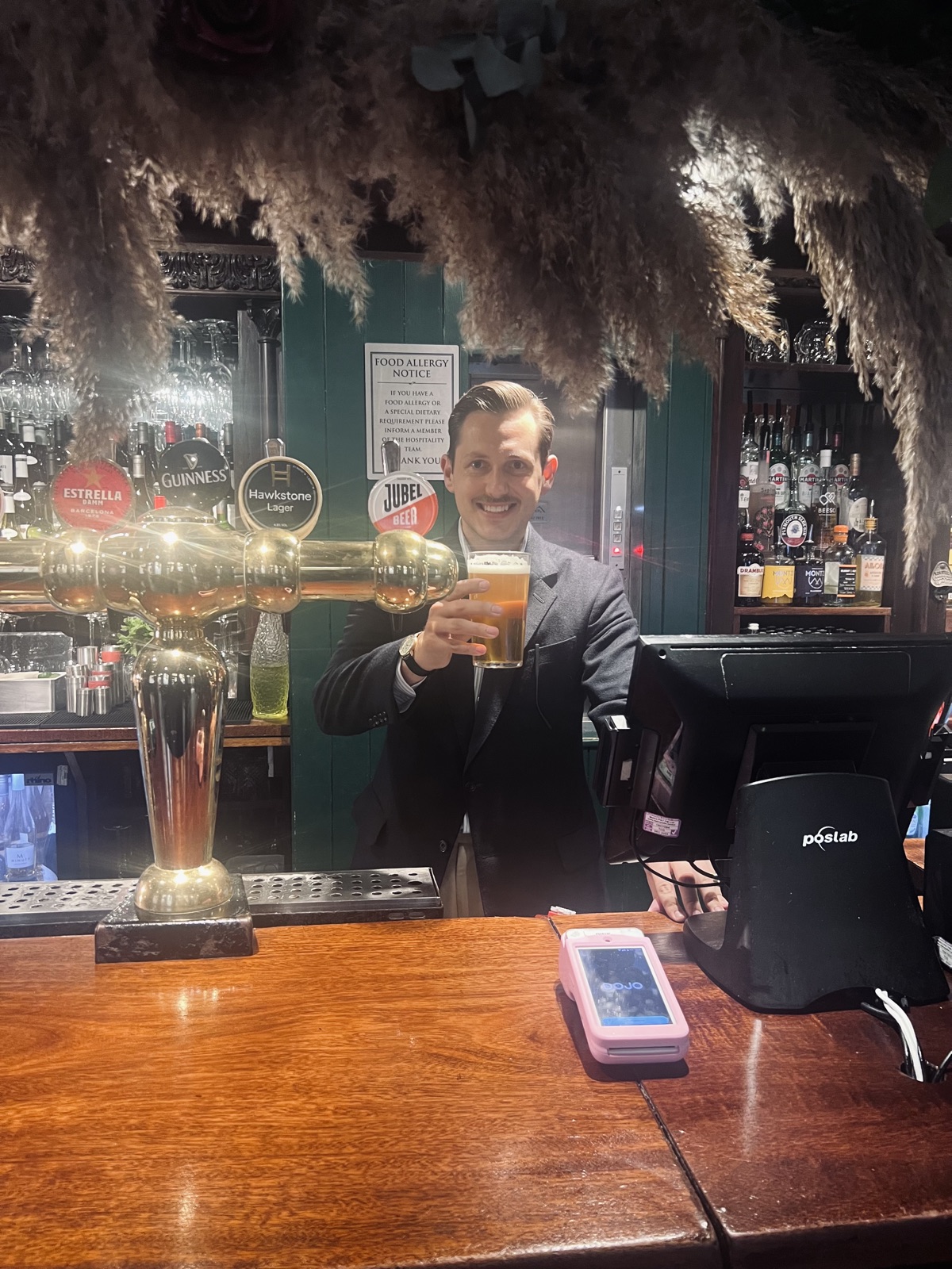 Behind the bar at the Lamb Tavern, Liam Randall in a dark jacket raises a freshly pulled pint of golden ale above the polished brass font, its badges for Estrella, Guinness, Hawkstone and Jubel; green panelling, spirit shelves and hanging pampas grass fill the bar behind him, and a bright pink card reader sits propped on the counter beside the till.