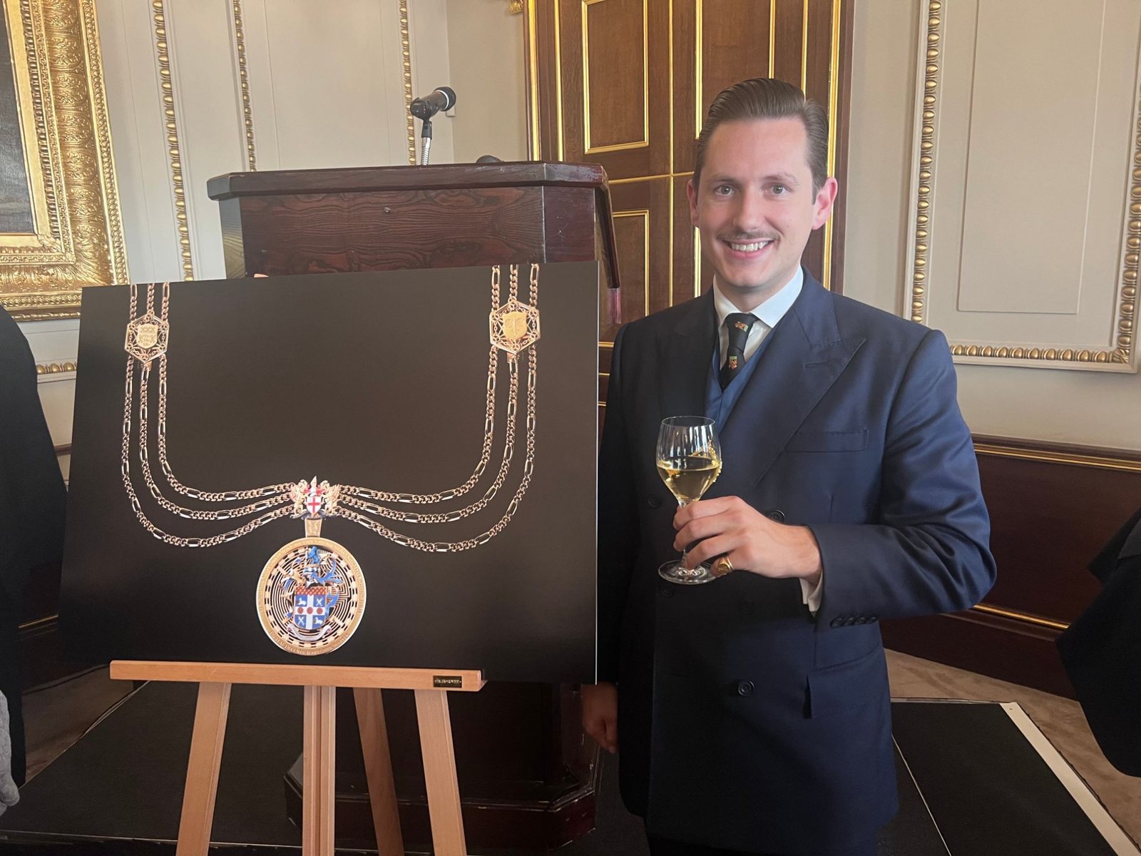 Liam Randall stands beside an easel displaying an image of the Sheriff&rsquo;s new chain of office and badge at Fishmongers&rsquo; Hall.