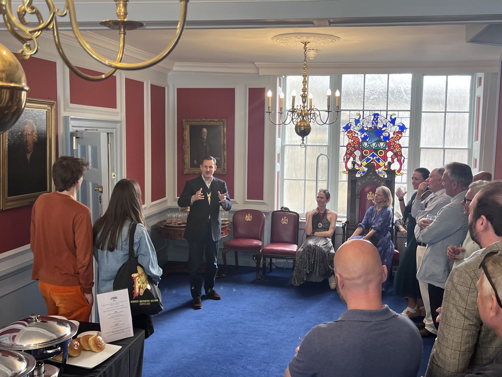 A tasting under way in a panelled room at Coopers&rsquo; Hall: deep red walls, a blue carpet and brass chandeliers, with Liam Randall standing mid-sentence before a semicircle of guests. Tasting glasses wait on a side table and the Company&rsquo;s coat of arms glows in stained glass in the bay window behind him.
