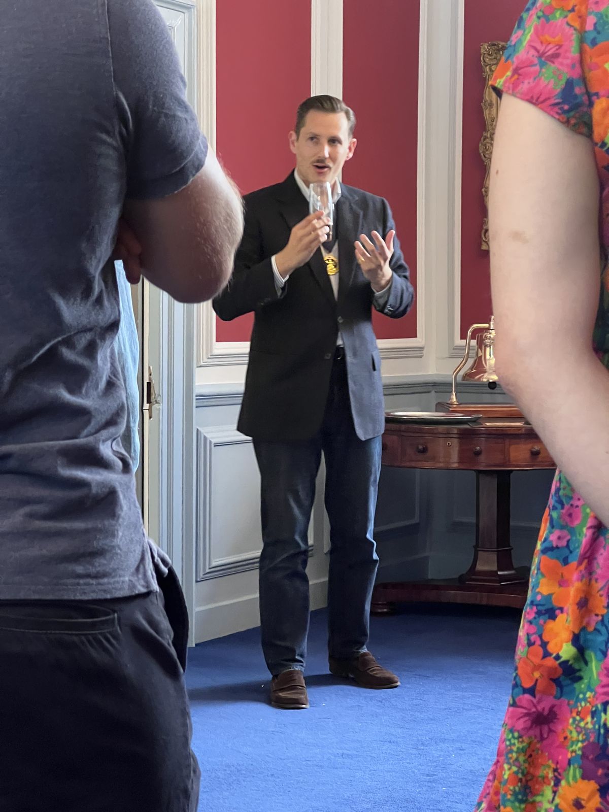 Liam Randall in a dark jacket with the Ale-Conner&rsquo;s badge of office at his chest, holding a tasting glass as he speaks, framed between two guests standing in the foreground.