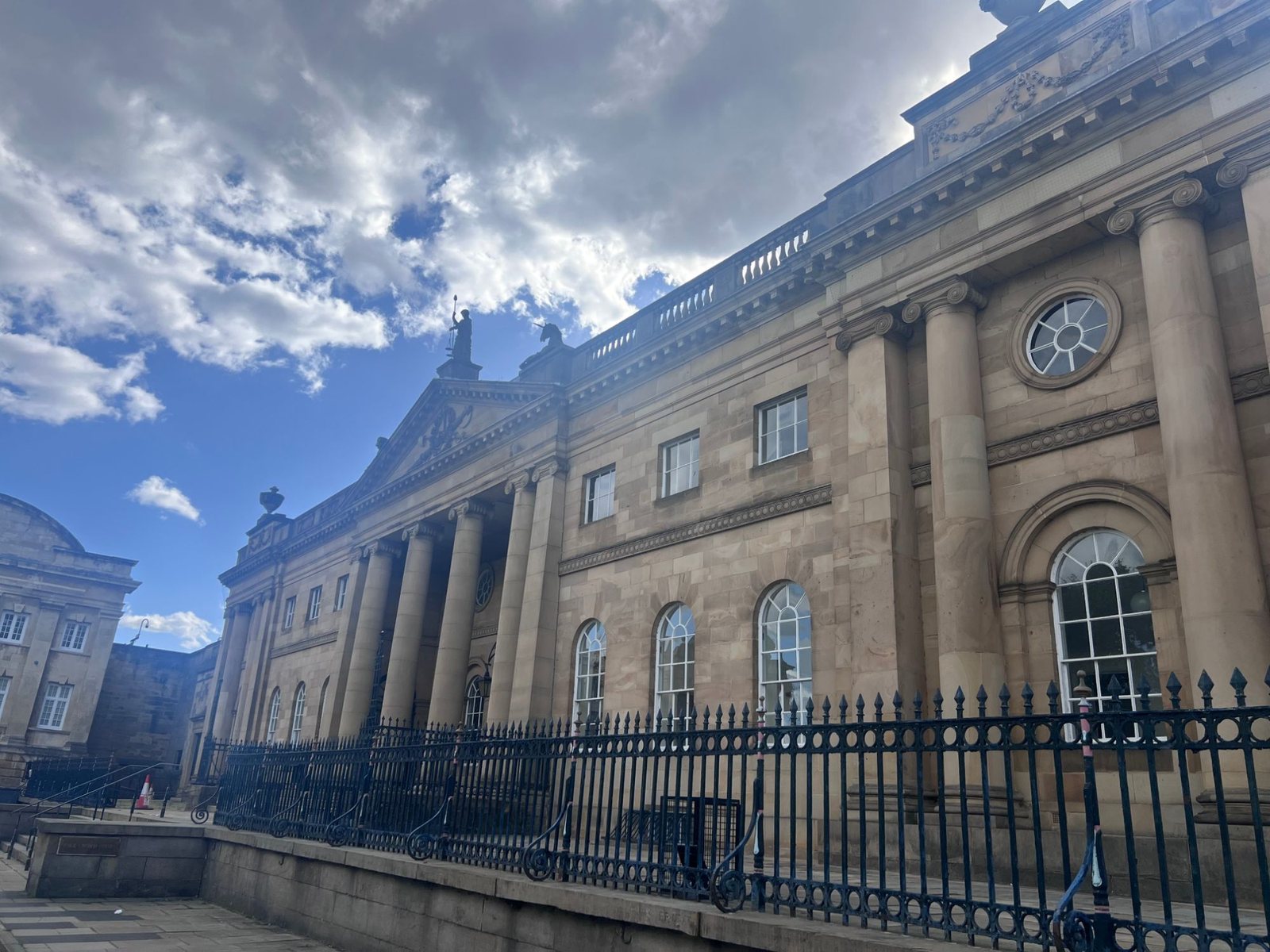The Georgian stone front of York Crown Court under a bright, cloudy sky, its columned portico topped by a statue of Justice and a unicorn, with black iron railings along the pavement.